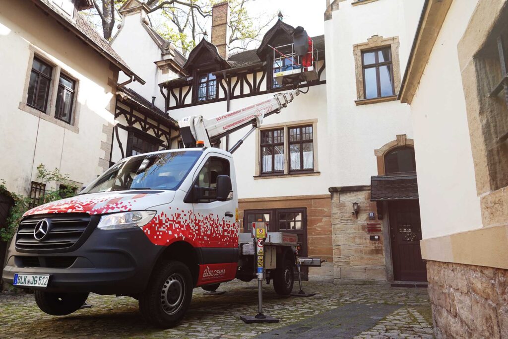 Ein Firmenfahrzeug von Bloehm Dachrinnenreinigung mit Arbeitsbühne steht in einem Innenhof. Über die ausgefahrene Hebebühne reinigt ein Mitarbeiter die Dachrinne eines Gebäudes.
